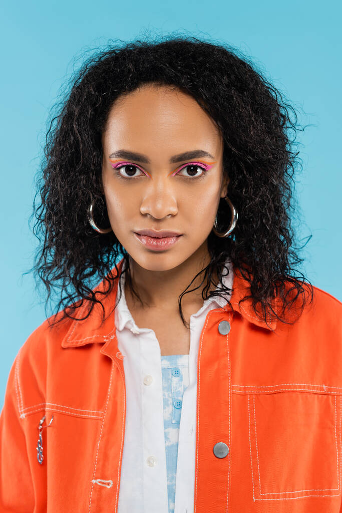Porträt einer afrikanisch-amerikanischen Frau mit welligem Haar und hellem Make-up, die isoliert auf blauem Grund in die Kamera blickt - Foto, Bild