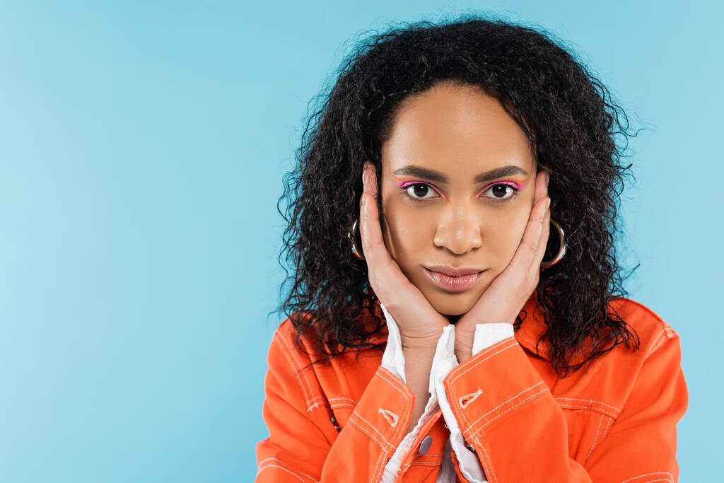 portrét brunetky africké americké ženy v oranžové bundě dotýkající se obličeje a při pohledu na kameru izolované na modré - Fotografie, Obrázek