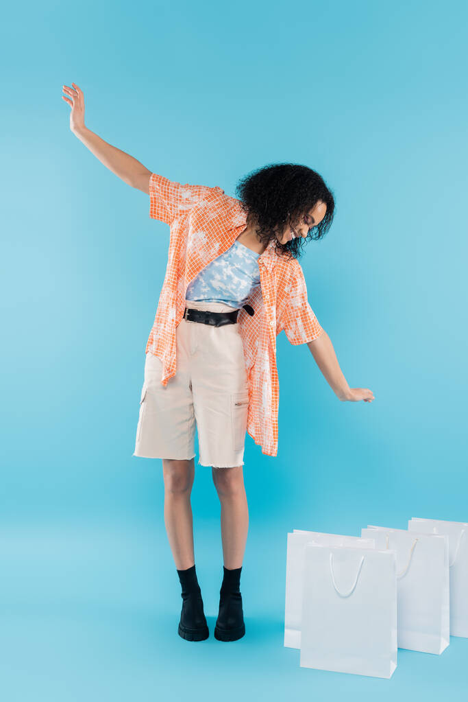 comprimento total de mulher americana africana feliz em botas pretas e shorts olhando para sacos de compras brancos no fundo azul - Foto, Imagem