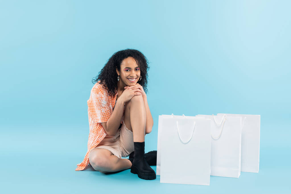 plná délka usmívající se africké americké ženy v módní oblečení sedí v blízkosti bílých nákupních tašek na modrém pozadí - Fotografie, Obrázek