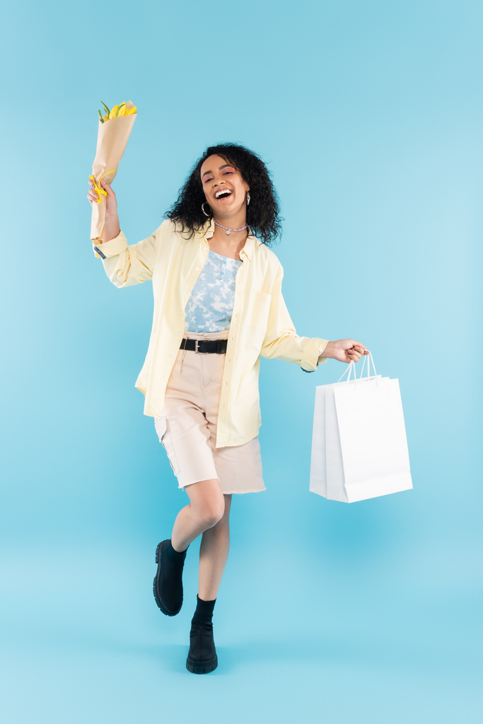volle Länge der überglücklichen afrikanisch-amerikanischen Frau in kurzen Hosen und schwarzen Stiefeln posiert mit gelben Tulpen und Einkaufstaschen auf blau - Foto, Bild