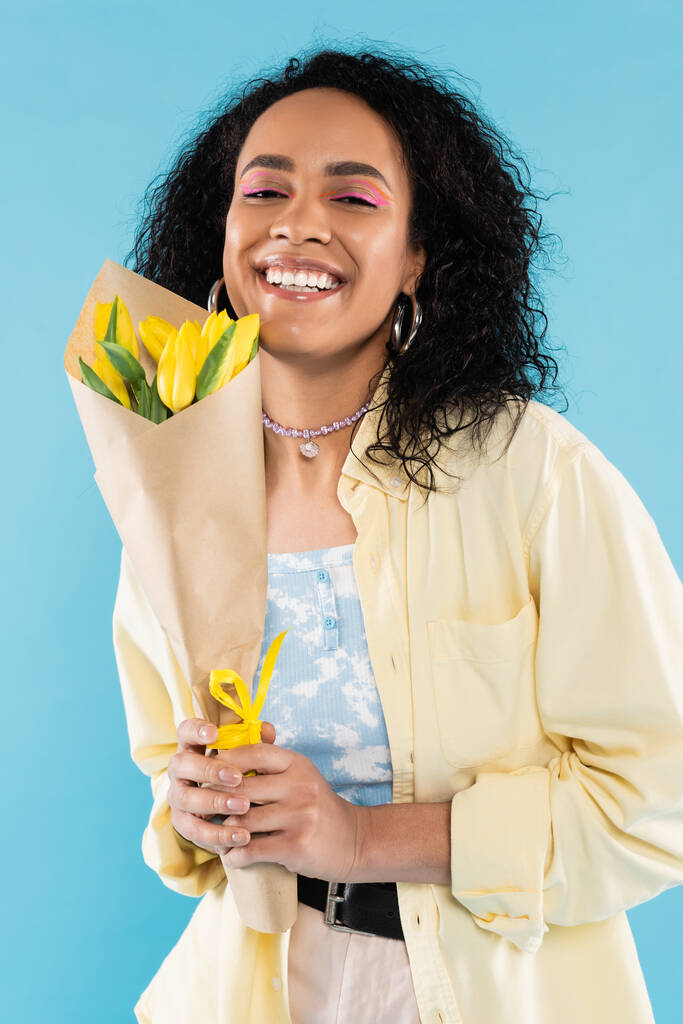 donna afroamericana trendy e felice con capelli ricci bruna che tengono tulipani gialli e guardando la fotocamera isolata sul blu - Foto, immagini