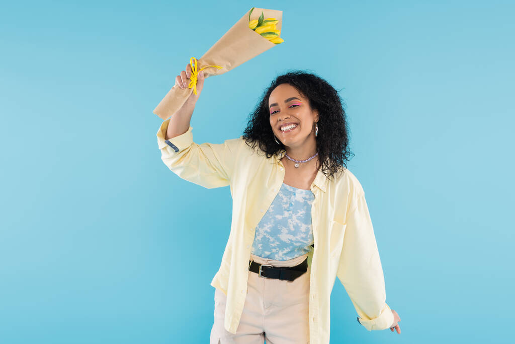 radosna i modna Afroamerykanka uśmiecha się do kamery trzymając żółte tulipany odizolowane na niebiesko - Zdjęcie, obraz