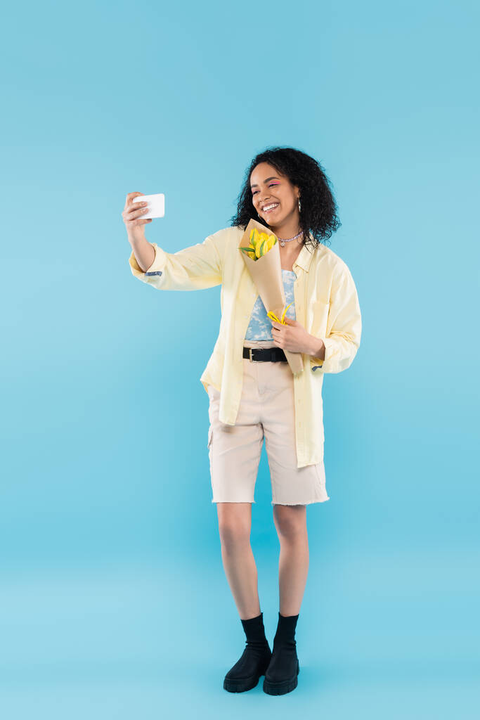 longitud completa de la sonriente mujer afroamericana con tulipanes amarillos tomando selfie en el teléfono móvil en azul - Foto, imagen