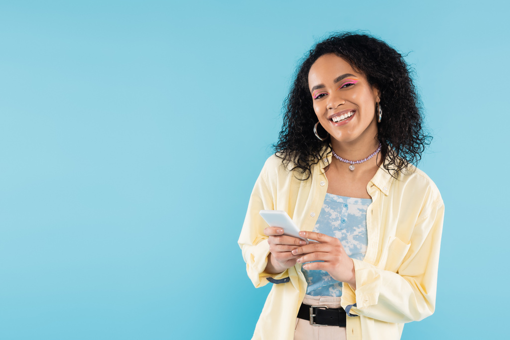 χαρούμενη Αφροαμερικανή γυναίκα με κομψά ρούχα που κρατάει smartphone και κοιτάζει την κάμερα απομονωμένη στο μπλε - Φωτογραφία, εικόνα
