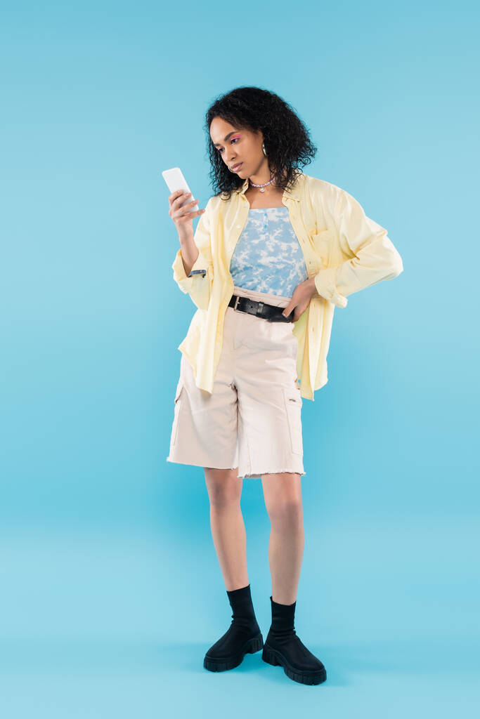 longitud completa de la mujer afroamericana con estilo en pantalones cortos y botas negras mirando el teléfono inteligente sobre fondo azul - Foto, Imagen
