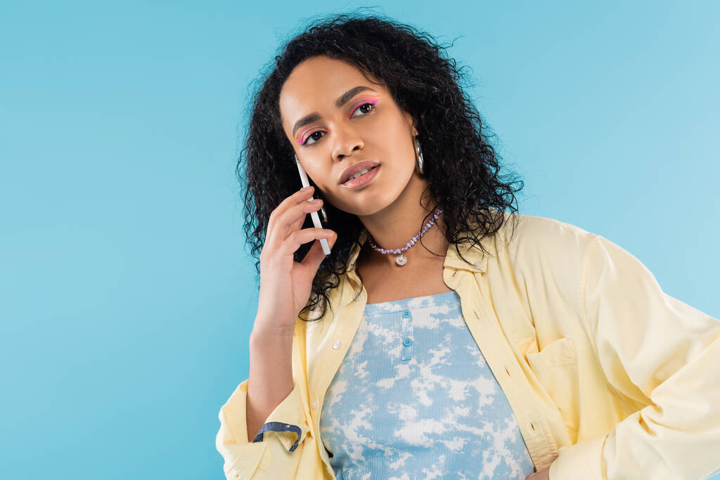 morena afro-americana em camisa amarela falando no celular e olhando para longe isolado no azul - Foto, Imagem