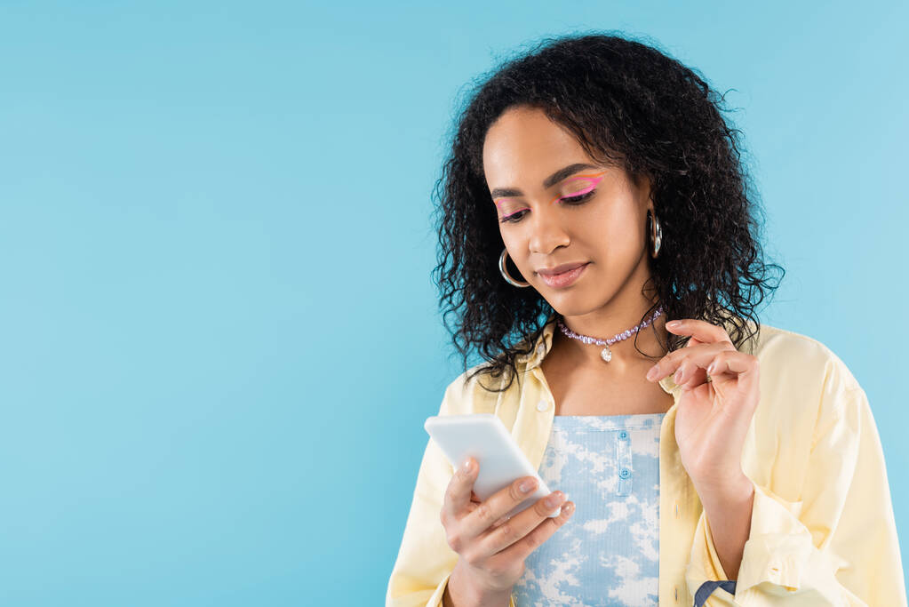stylowa Afroamerykanka w obręczy kolczyki i koraliki za pomocą telefonu komórkowego odizolowany na niebiesko - Zdjęcie, obraz