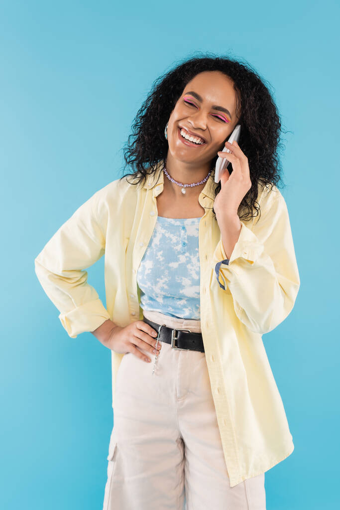 mujer afroamericana de moda y feliz de pie con la mano en la cadera y hablando en el teléfono celular aislado en azul - Foto, imagen