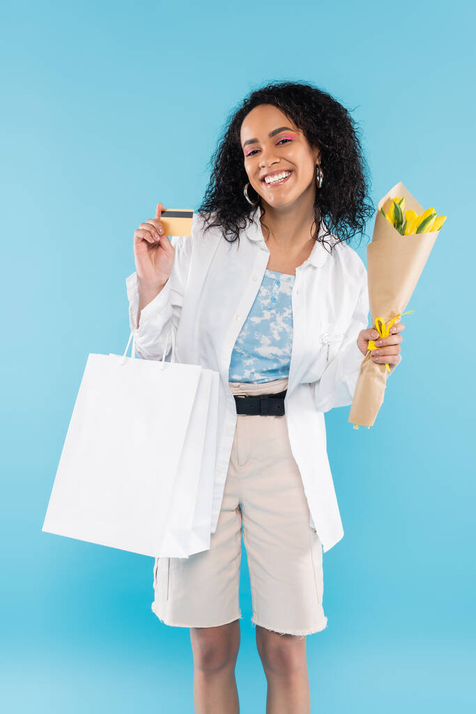 vrolijke Afrikaans-Amerikaanse vrouw met boodschappentassen en gele tulpen tonen credit card geïsoleerd op blauw - Foto, afbeelding