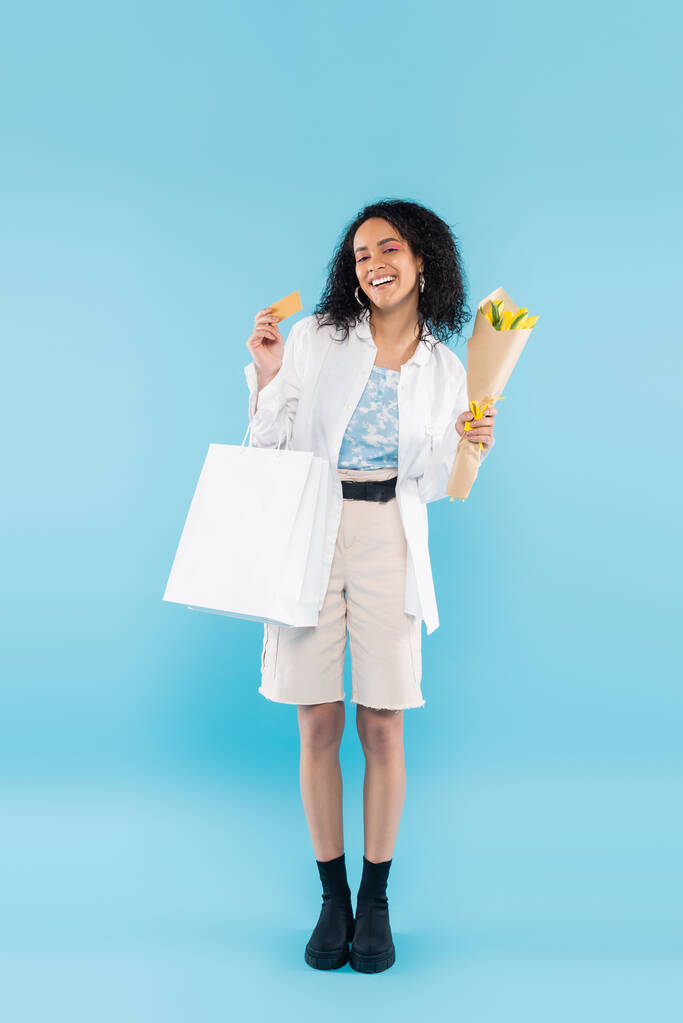 青い背景にクレジットカードを示す黄色のチューリップやショッピングバッグを持つ陽気なアフリカ系アメリカ人女性の完全な長さ - 写真・画像