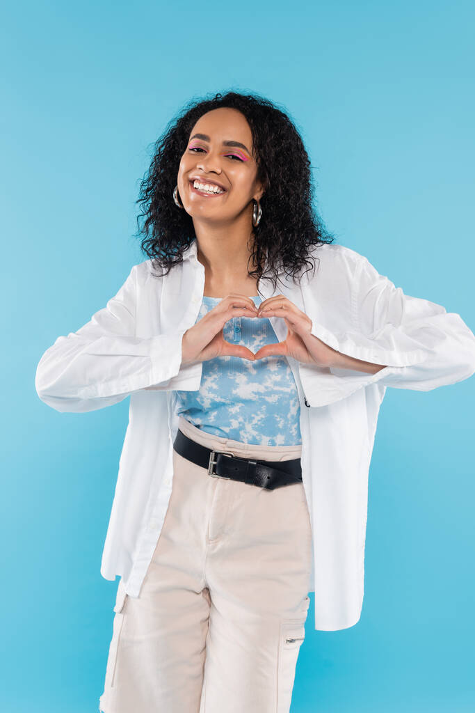 ξέγνοιαστη αφροαμερικανίδα με λευκό κομψό πουκάμισο που δείχνει το σήμα της καρδιάς με τα χέρια απομονωμένα στο μπλε - Φωτογραφία, εικόνα