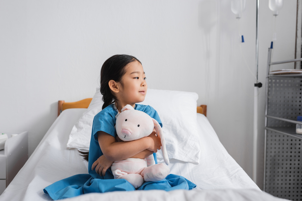 pensive asian girl hugging toy bunny and looking away while sitting on hospital bed - Photo, Image