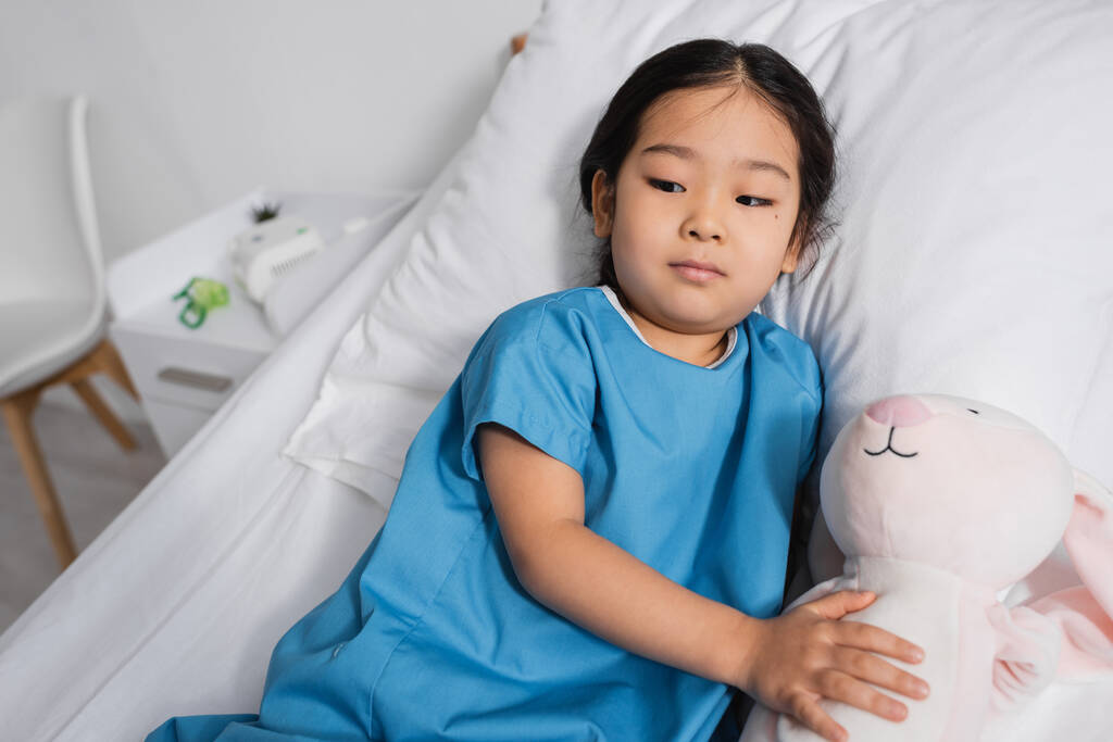 coûteux asiatique enfant couché sur lit d'hôpital avec jouet lapin et regarder loin - Photo, image