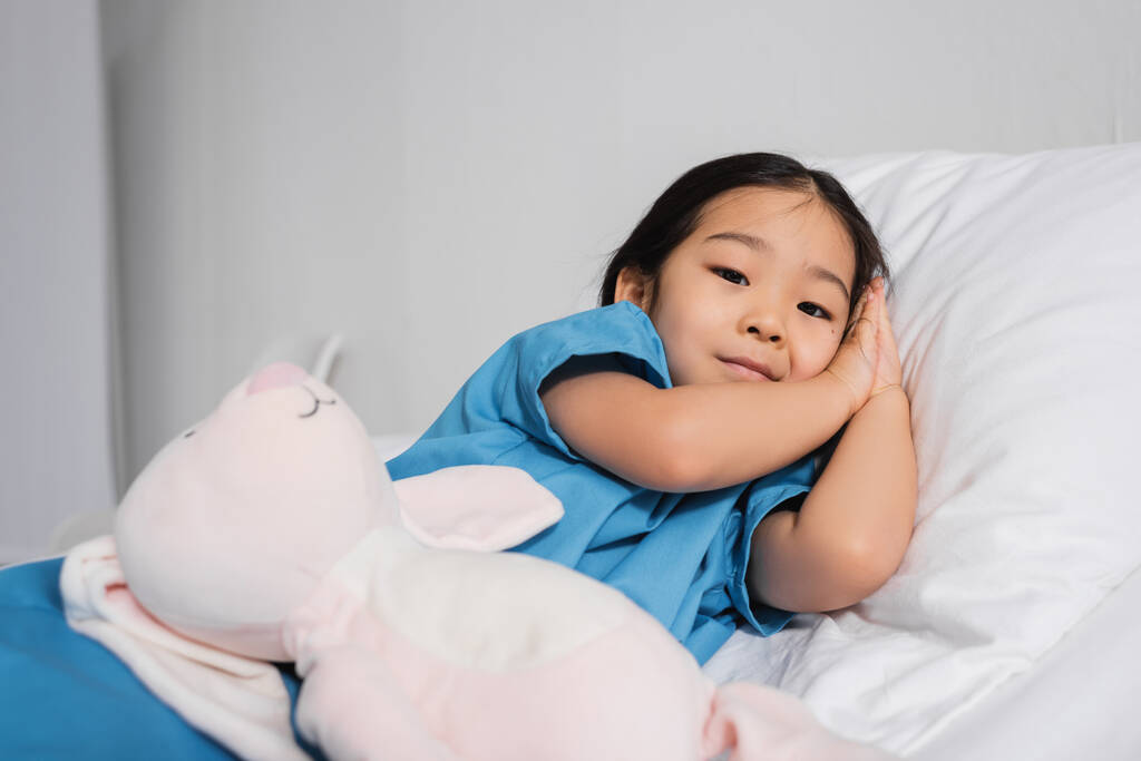 θετική ασιατική παιδί κοιτάζοντας κάμερα, ενώ βρίσκεται στο κρεβάτι του νοσοκομείου κοντά παιχνίδι λαγουδάκι - Φωτογραφία, εικόνα