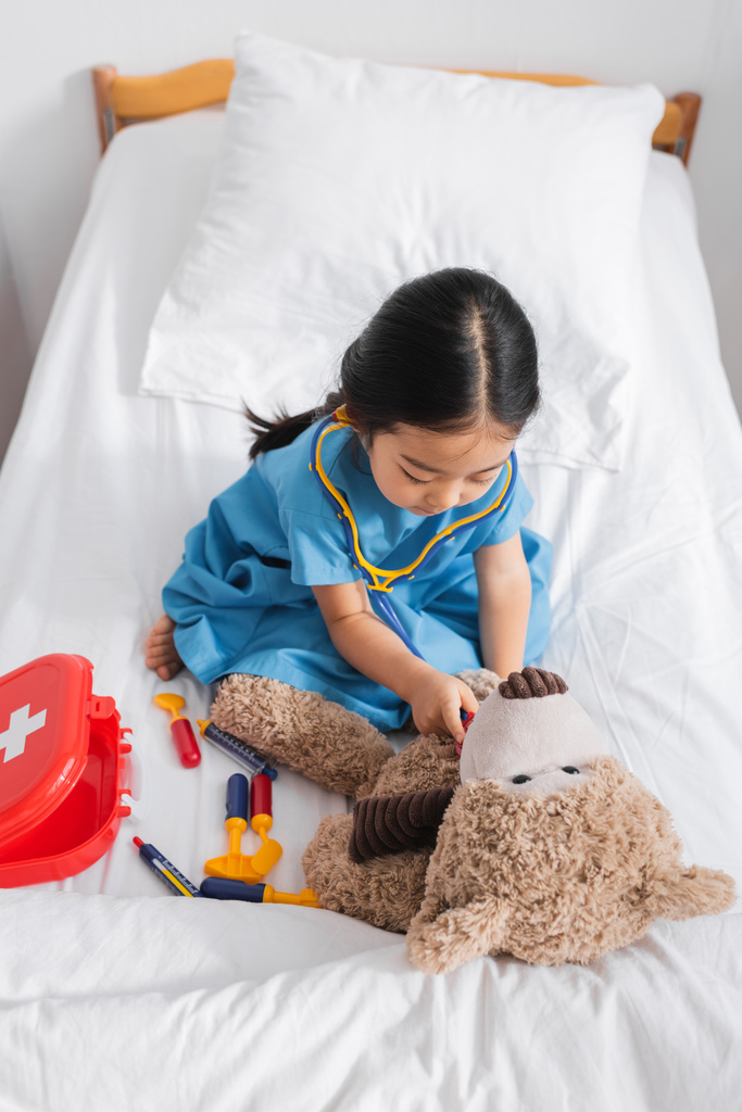 alto ângulo vista de ásia menina examinando ursinho de pelúcia com brinquedo estetoscópio enquanto jogar no cama no hospital - Foto, Imagem