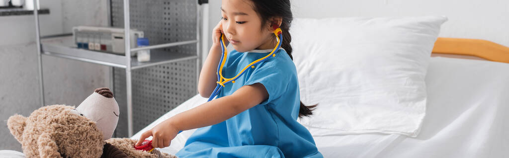 Hastane yatağında oyuncak steteskopla oyuncak ayıyı muayene eden Asyalı kız. - Fotoğraf, Görsel