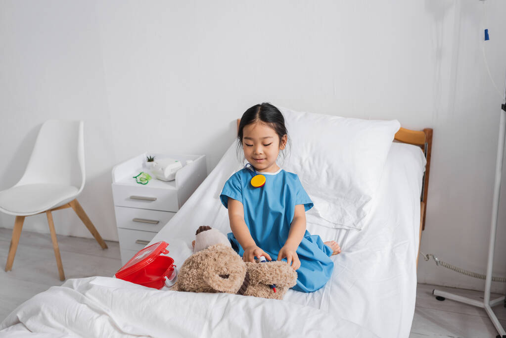 Çocuk kliniğinde oyuncak ayı ve oyuncak tıbbi malzemelerle oynayan neşeli Asyalı çocuk. - Fotoğraf, Görsel