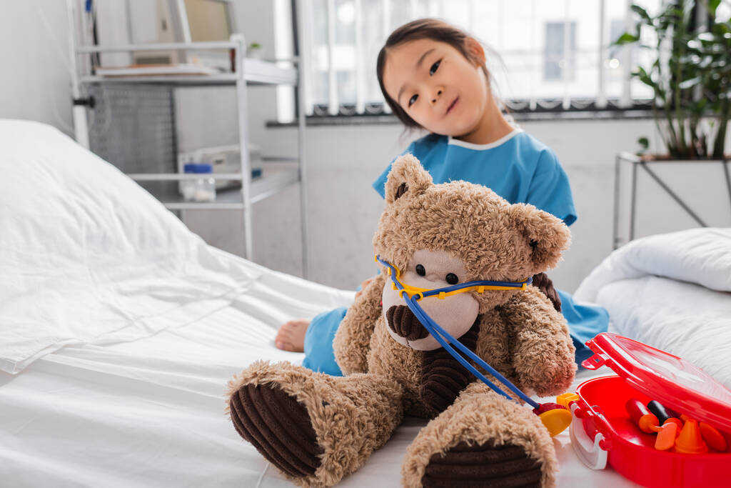 Ασιάτισσα που κοιτάει την κάμερα ενώ παίζει με αρκουδάκι και παιχνίδια ιατρικού εξοπλισμού στο κρεβάτι του νοσοκομείου - Φωτογραφία, εικόνα
