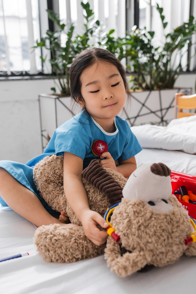 mosolygó ázsiai lány kórházi köpenyben játszik Teddy macival és játék sztetoszkóppal az ágyon a gyermekklinikán - Fotó, kép