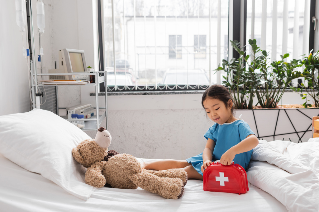 asiático chica apertura juguete primeros auxilios kit cerca teddy oso en cama en moderno pediátrico clínica - Foto, imagen