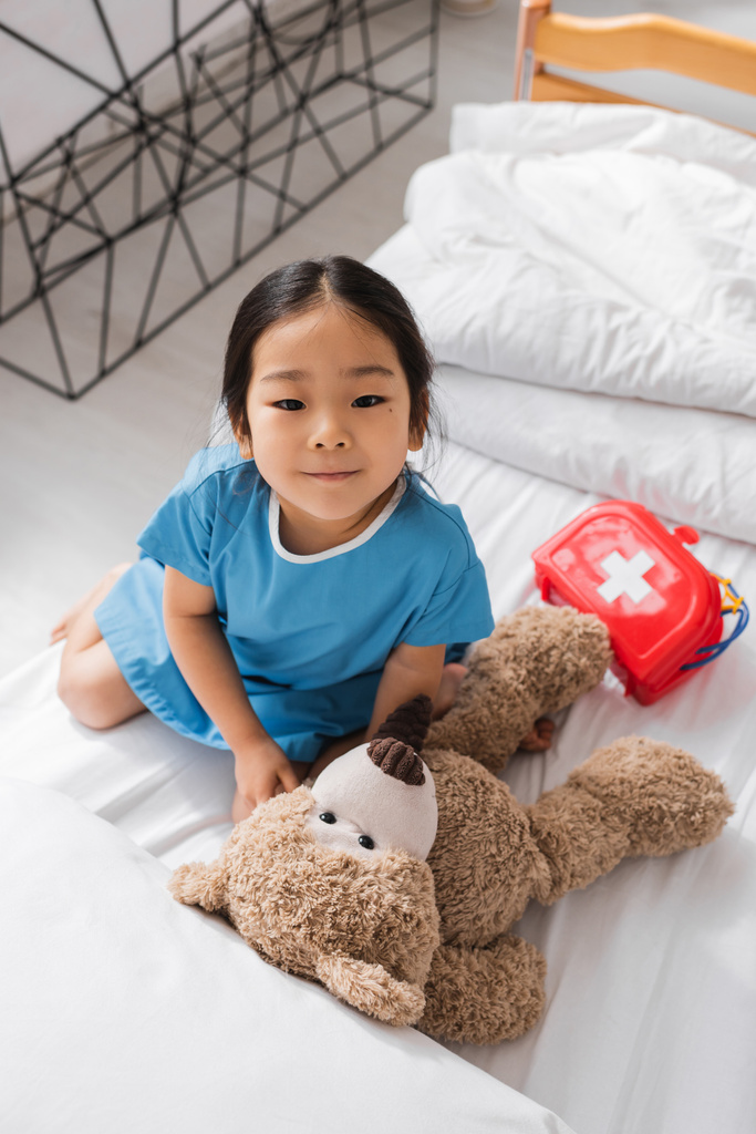 вид сверху на азиатскую девушку, улыбающуюся в камеру во время игры с плюшевым мишкой и игрушечным медицинским оборудованием на больничной койке - Фото, изображение