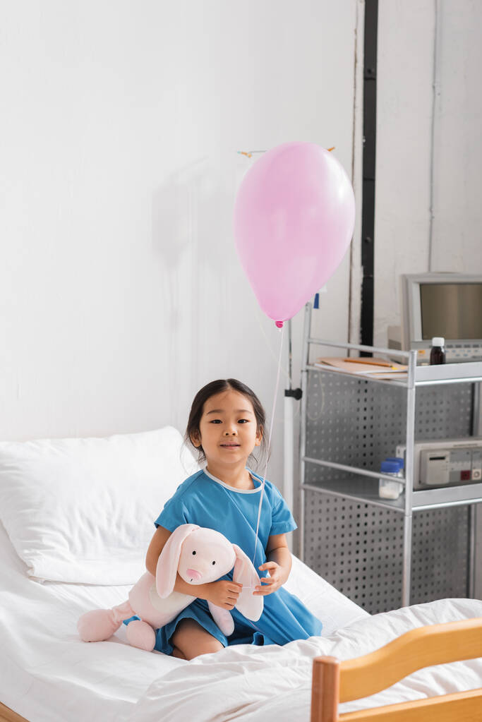 беззаботная азиатская девушка улыбается в камеру, сидя с воздушным шаром и игрушечным кроликом на больничной койке - Фото, изображение