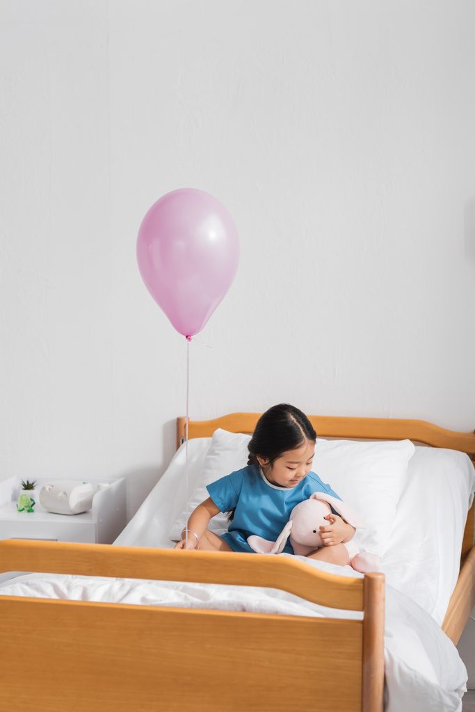 Asijské dítě v nemocniční šaty sedí na posteli s měkkou hračkou pod slavnostní balón - Fotografie, Obrázek