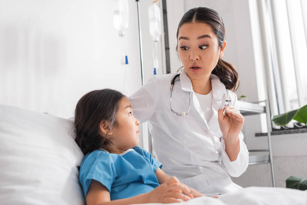 asiatischer Arzt zeigt Stethoskop und spricht mit kleinem Patienten in Kinderklinik - Foto, Bild