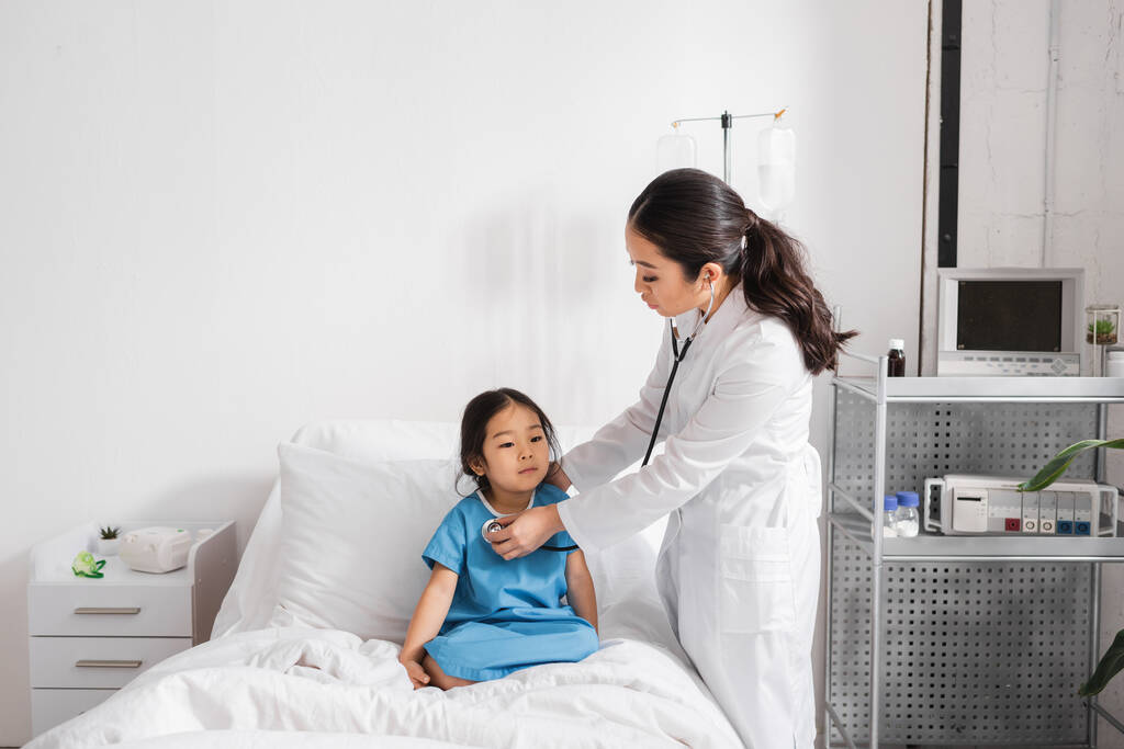 педиатр в белом халате осматривает азиатку со стетоскопом в больничном отделении - Фото, изображение