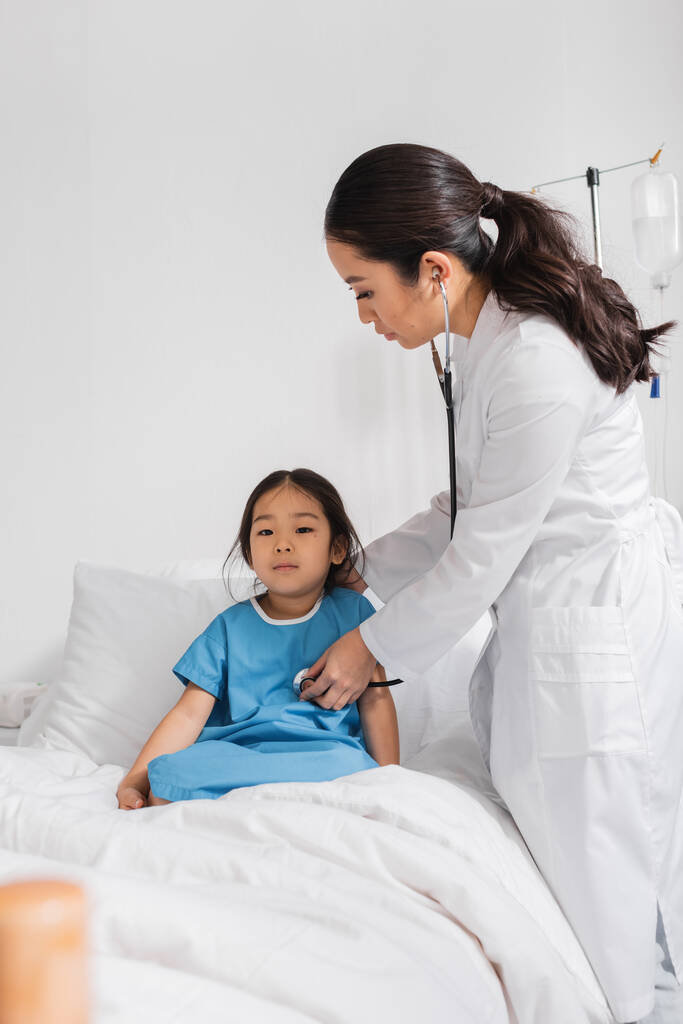 Junge Kinderärztin untersucht asiatisches Kind mit Stethoskop auf Bett in Klinik - Foto, Bild
