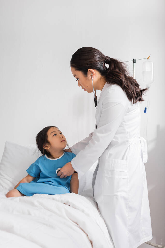 Asijské dítě sedí na nemocniční posteli a dívá se na pediatra se stetoskopem - Fotografie, Obrázek