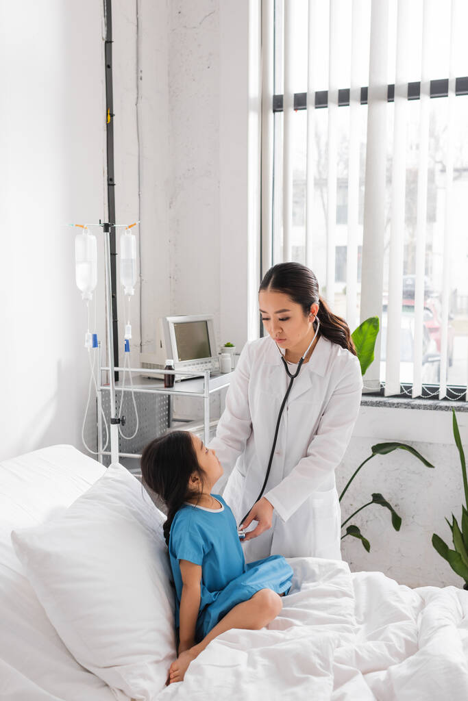 азиатский педиатр со стетоскопом осматривает маленького пациента, сидящего на больничной койке - Фото, изображение