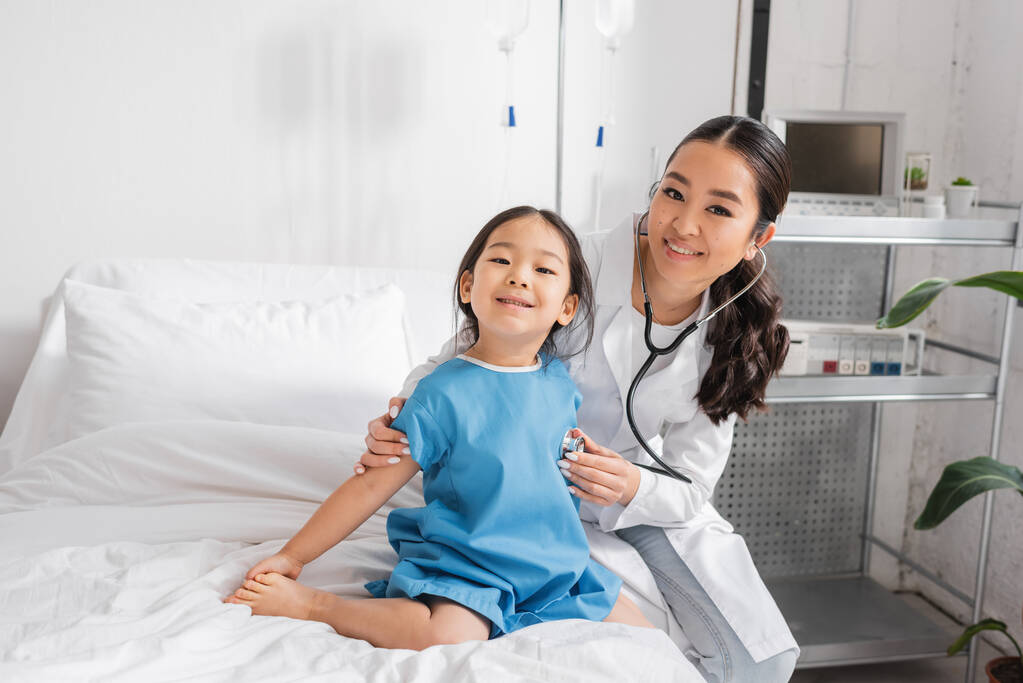 hymyilevä aasialainen lääkäri stetoskoopilla katsoen kameraa tutkiessaan pikkutyttöä lastenklinikalla - Valokuva, kuva