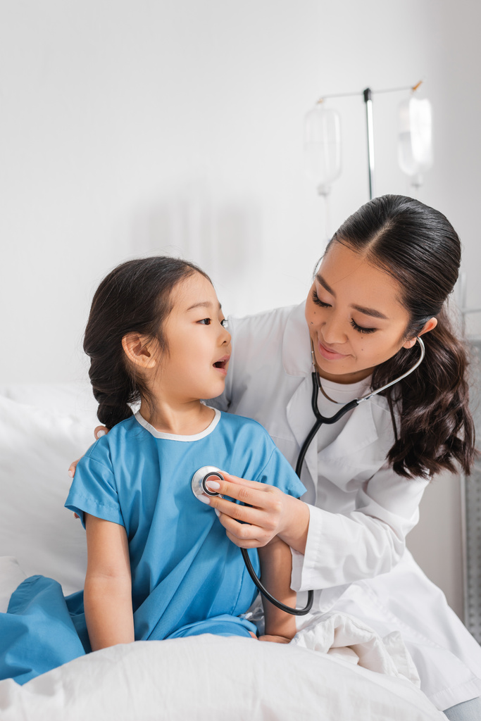 азіатська дівчина розмовляє зі усміхненим педіатром зі стетоскопом у лікарняному палаті
 - Фото, зображення