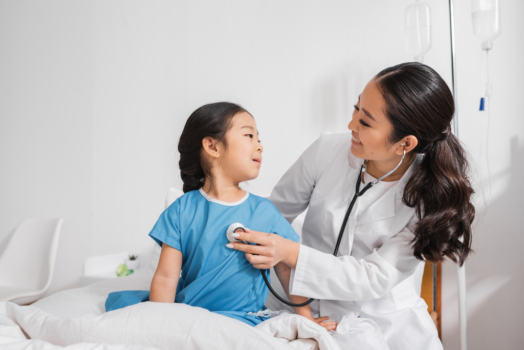 médico positivo em casaco branco examinando menina asiática com estetoscópio em hospital ward - Foto, Imagem