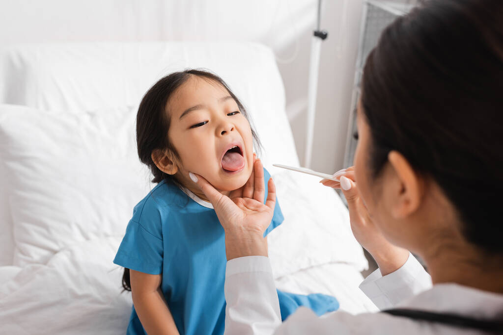 sfocato pediatra holding lingua depressore vicino asiatico ragazza apertura bocca in clinica - Foto, immagini