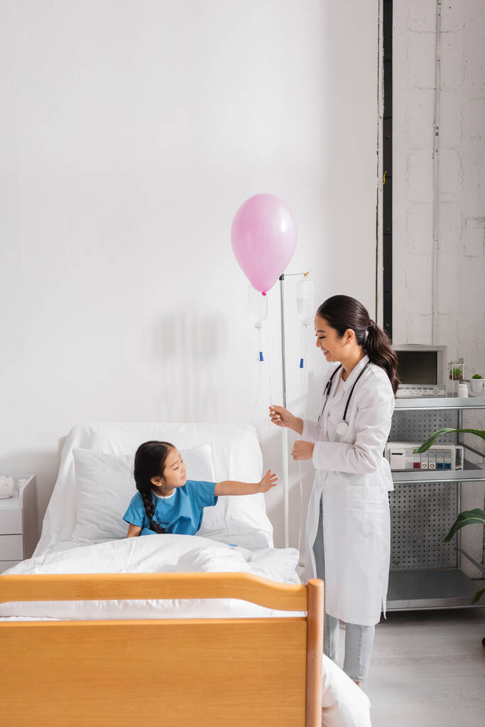 χαμογελαστός γιατρός με λευκό παλτό στέκεται με εορταστικό μπαλόνι κοντά στο ασιατικό κορίτσι στο κρεβάτι του νοσοκομείου - Φωτογραφία, εικόνα