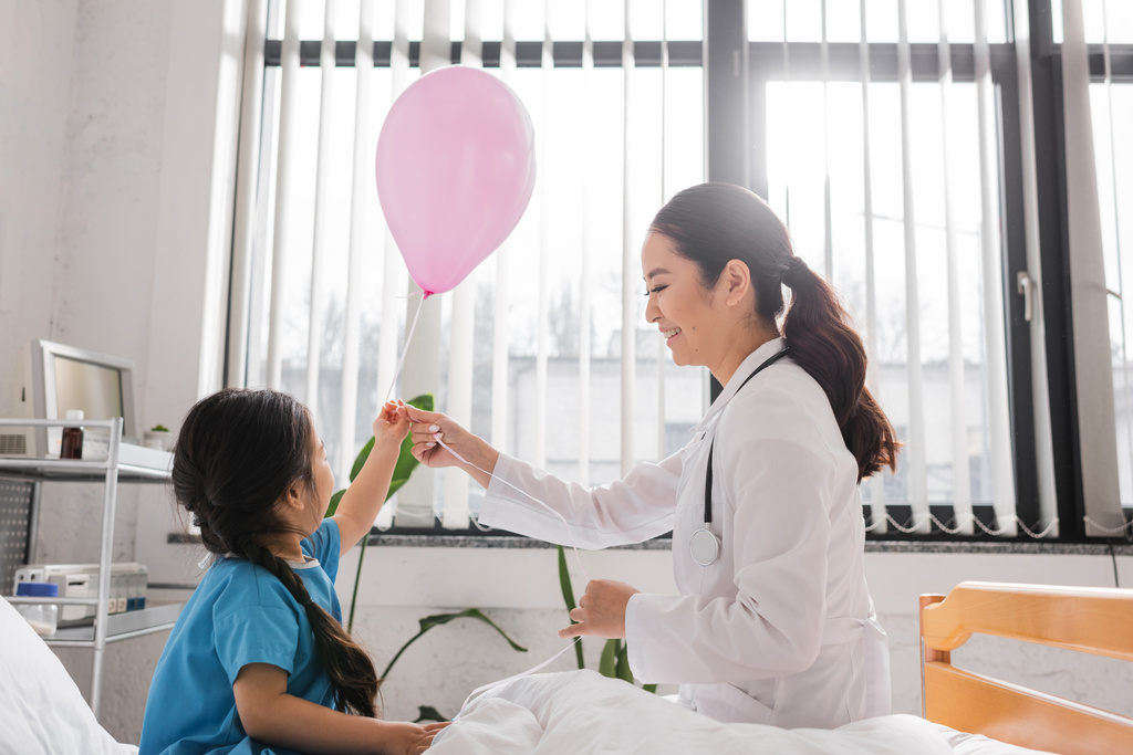 Hastanenin koğuşunda küçük bir kızın yanında elinde balon tutan mutlu Asyalı doktorun yan görüntüsü. - Fotoğraf, Görsel