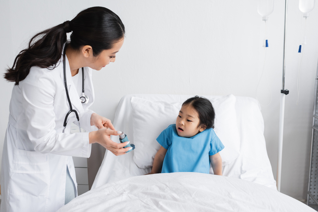 joven médico mostrando inhalador a sorprendido asiático chica sentado en cama en hospital sala - Foto, Imagen