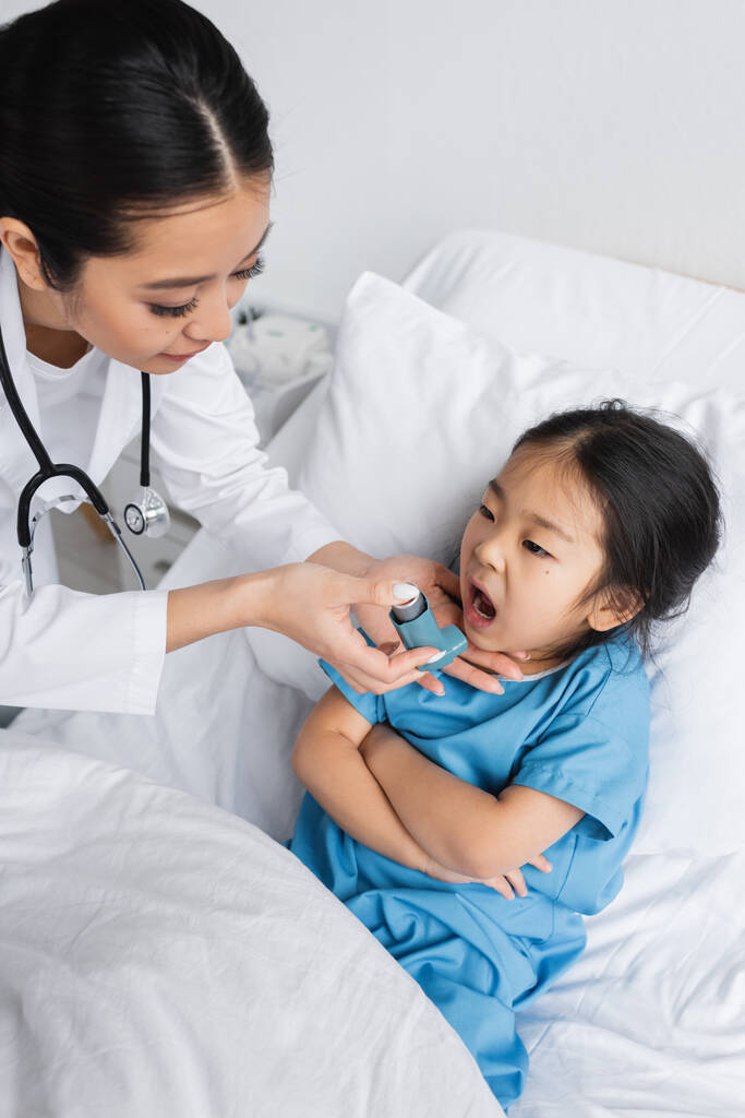 joven pediatra celebración inhalador cerca asiático chica con abierto boca y brazos cruzados en hospital cama - Foto, imagen