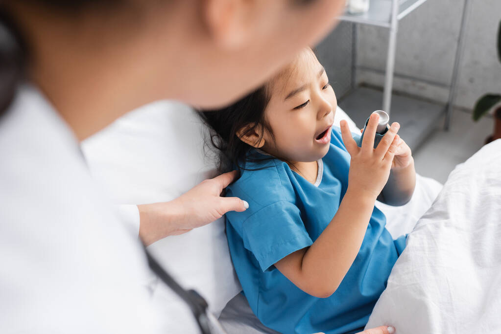 niewyraźny pediatra dotykający ramienia azjatyckiego dziecka stosującego inhalator na oddziale szpitalnym - Zdjęcie, obraz