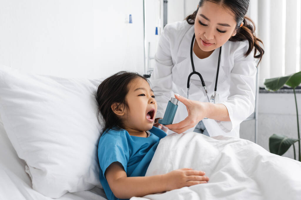 jeune asiatique pédiatre tenant inhalateur près de peu patient couché avec la bouche ouverte sur lit d'hôpital - Photo, image