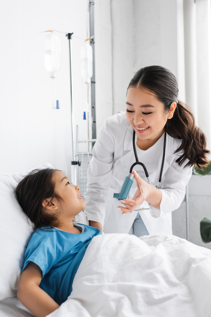 sonriente médico sosteniendo inhalador cerca positivo chica en cama en hospital sala - Foto, imagen