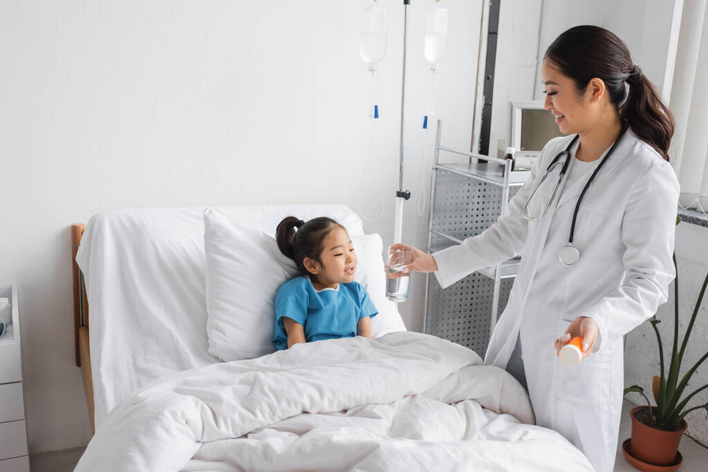 усміхнений педіатр тримає таблетки контейнер і склянку води біля безтурботної азіатської дівчини на ліжку в клініці
 - Фото, зображення