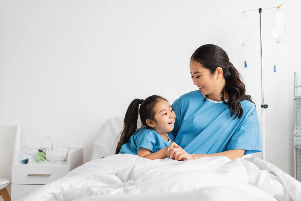病院病棟のベッドで笑顔の娘の手を握ってる若いアジア人女性 - 写真・画像
