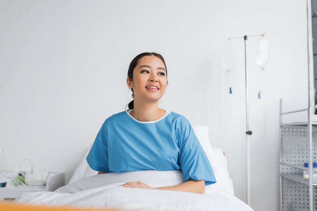 freudige asiatische Frau im Krankenhauskleid sitzt auf dem Bett in der Klinik und schaut weg - Foto, Bild