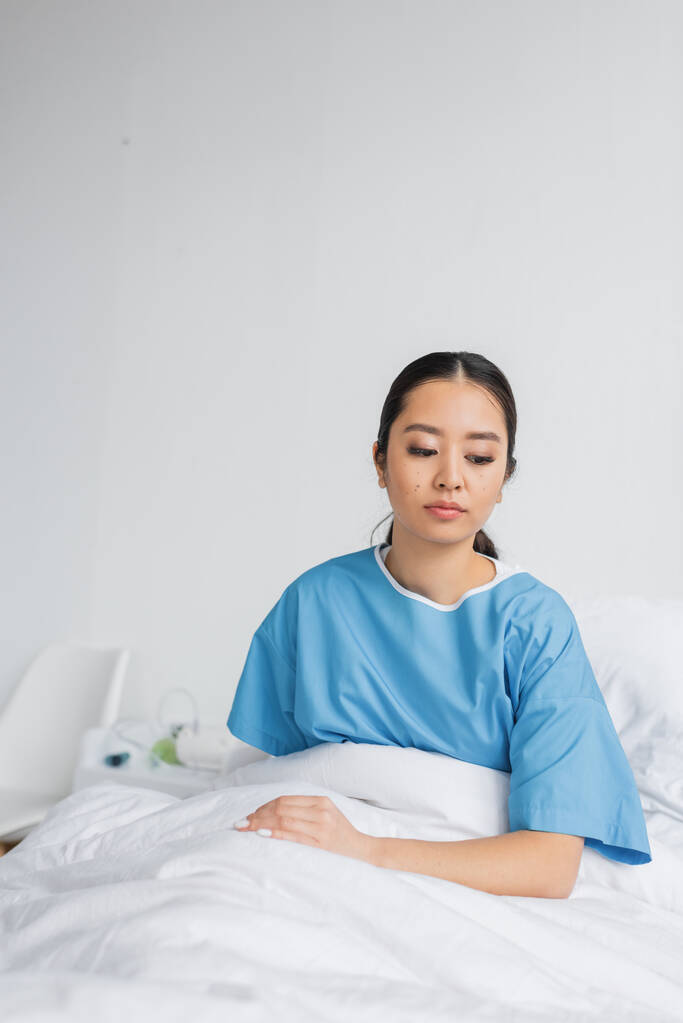 καταθλιπτική Ασιάτισσα γυναίκα με νοσοκομειακή ρόμπα σκέφτεται ενώ κάθεται στο κρεβάτι στην κλινική - Φωτογραφία, εικόνα
