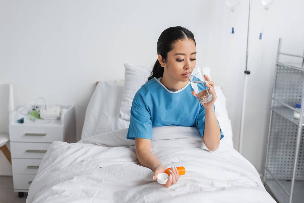 fiatal ázsiai nő ivóvíz és gazdaság tabletták tartály, miközben ül az ágyban a kórházi kórteremben - Fotó, kép