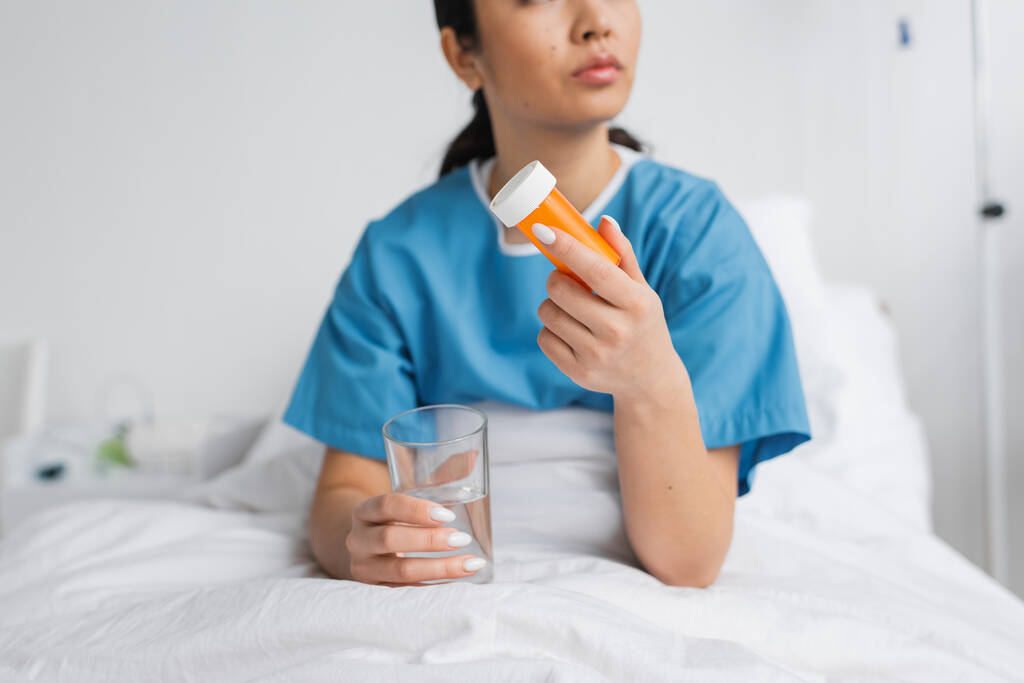 partial view of woman in hospital gown holding water and pills container on bed in clinic - Photo, Image